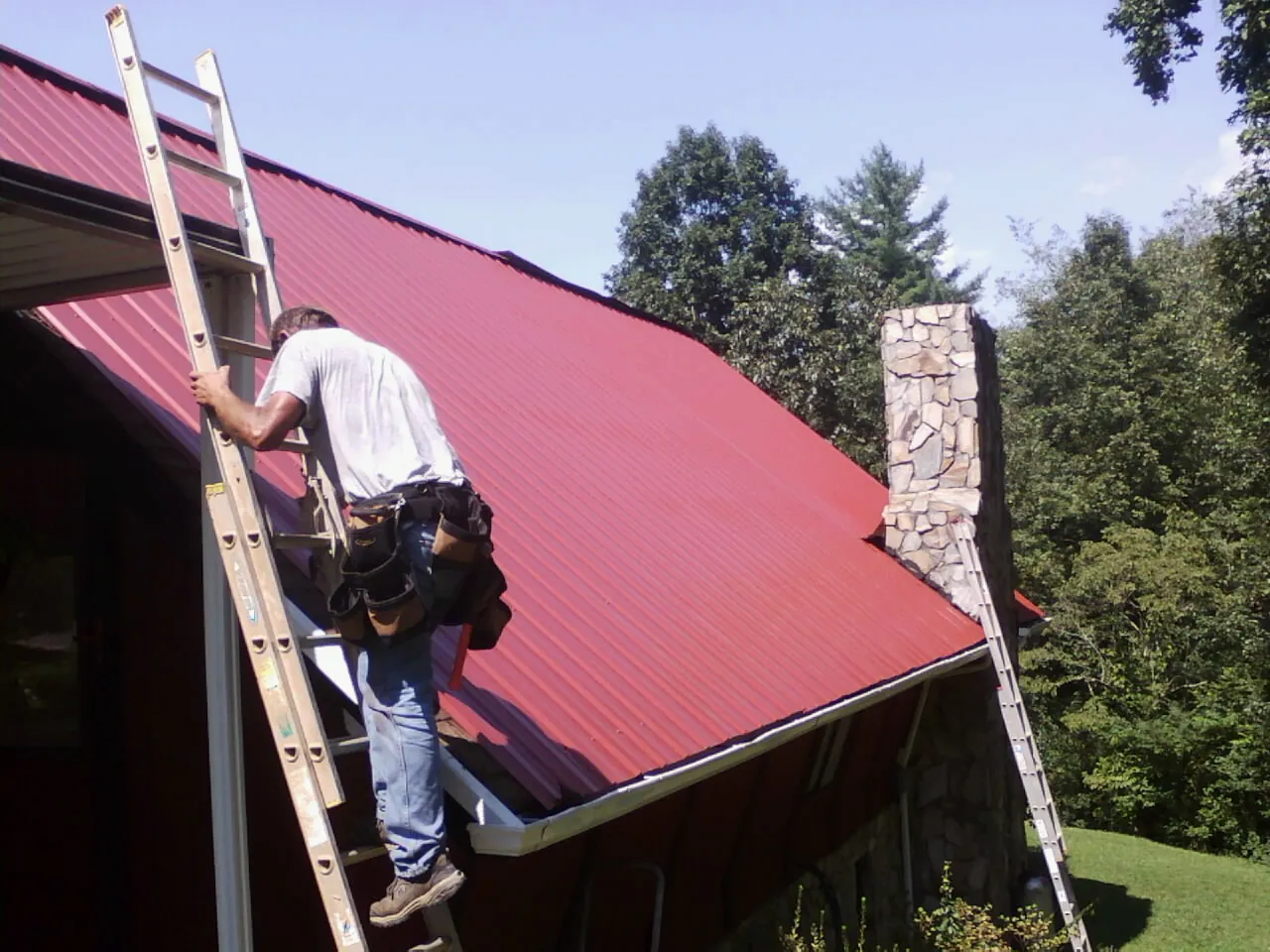 Roofer on a ladder beside a red metal roof and stone chimney — Soffit & Fascia Repair by Roofing Brothers in Hampden